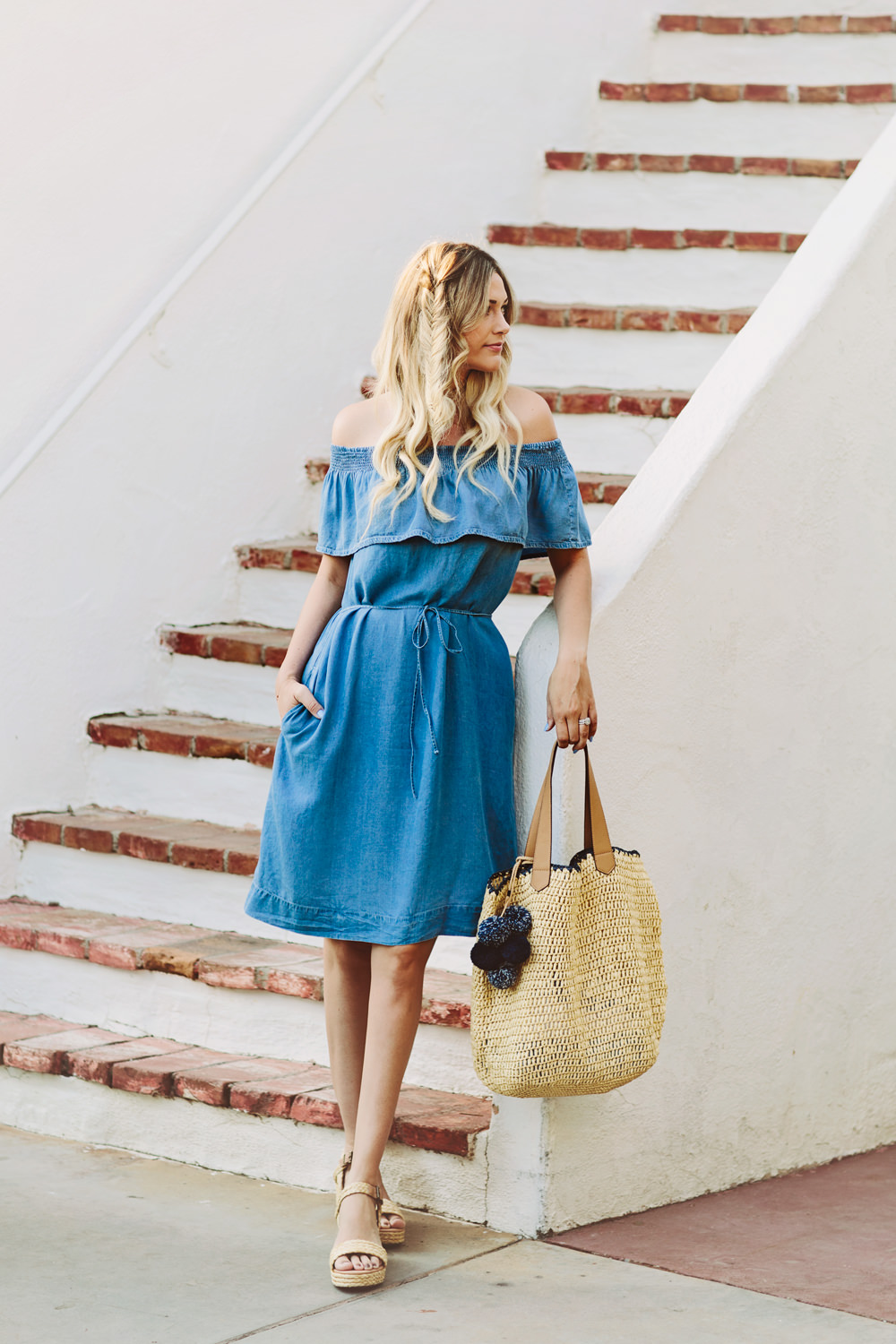 Caitlin Lindquist of Dash of Darling styles a chambray off shoulder dress from LOFT with a pom pom straw tote and wedge sandals for a summer look while in Palm Springs.