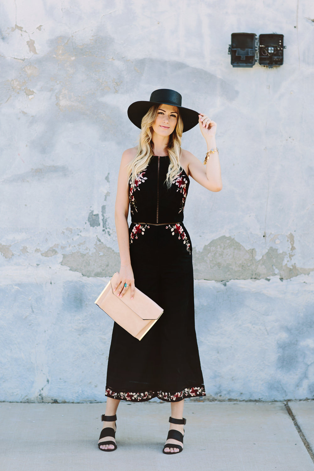 Caitlin Lindquist of Dash of Darling wears a Zimmermann floral embroidered black jumpsuit with a Janessa Leone straw hat and black sandals for a summer outfit perfect for a wedding.